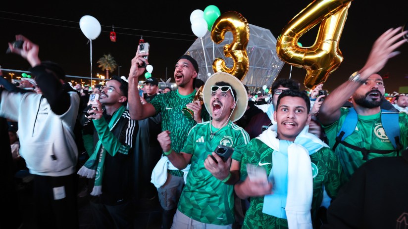 Excited crowd in green shirts and scarves cheering, holding phones and large gold balloons at night