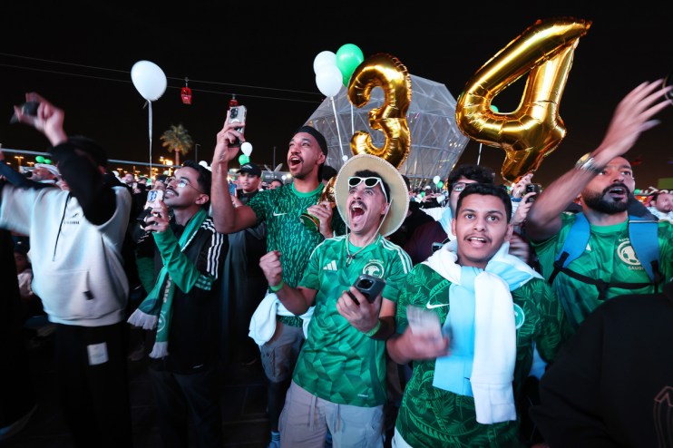 Excited crowd in green shirts and scarves cheering, holding phones and large gold balloons at night