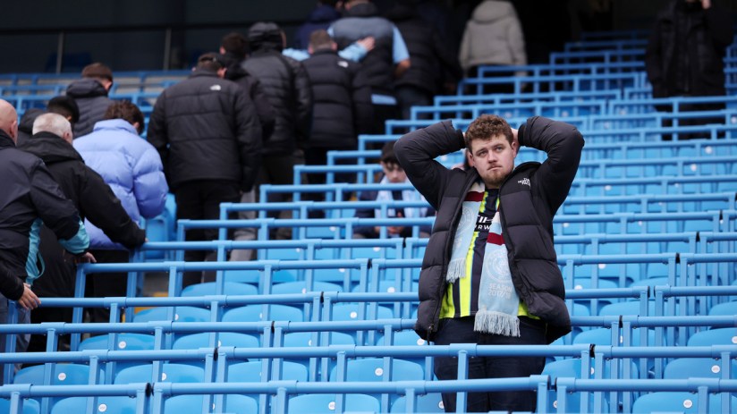 Disappointed football fan in a black puffer jacket and team scarf, hands on head, in a blue stadium crowd
