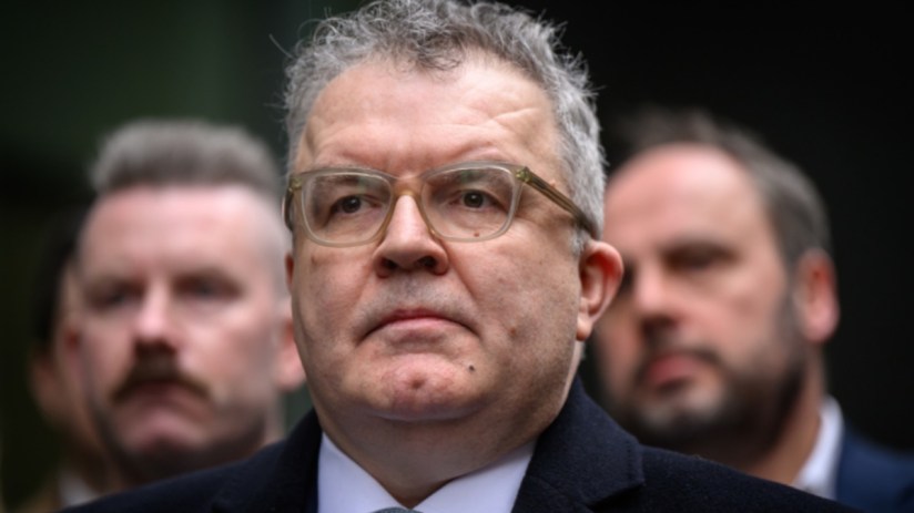 Tom Watson, former Labour Party Deputy Leader, wearing glasses and a dark coat, with two men blurred in the background.