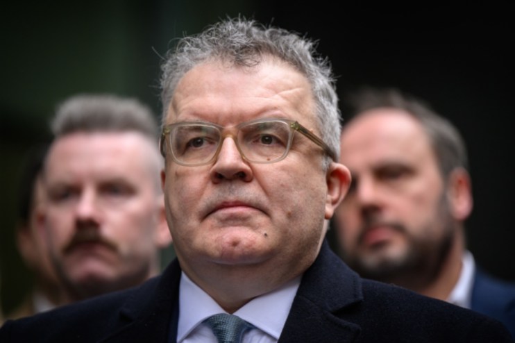 Tom Watson, former Labour Party Deputy Leader, wearing glasses and a dark coat, with two men blurred in the background.