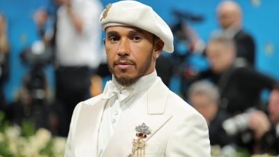 Lewis Hamilton in white suit, bow tie, beret, and pearl brooch looking directly at the camera.