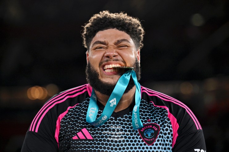 Soccer Aid player with beard bites gold medal, wearing black and pink kit, celebrating win for Unicef