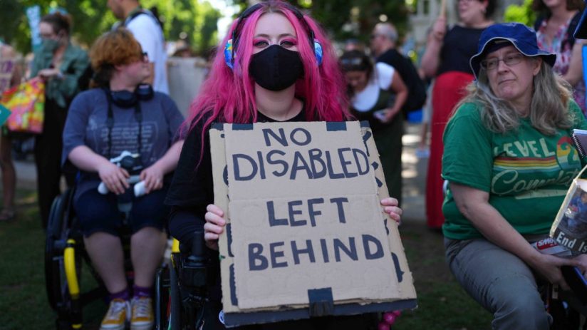 Person with pink hair in wheelchair holding NO DISABLED LEFT BEHIND sign at a protest