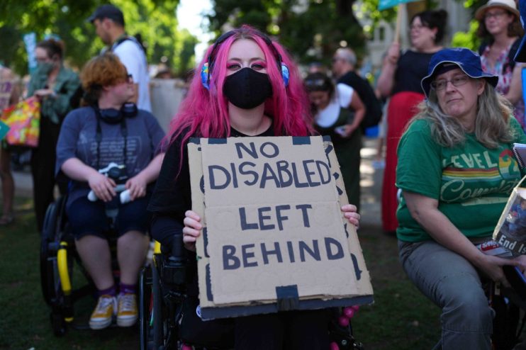 Person with pink hair in wheelchair holding NO DISABLED LEFT BEHIND sign at a protest