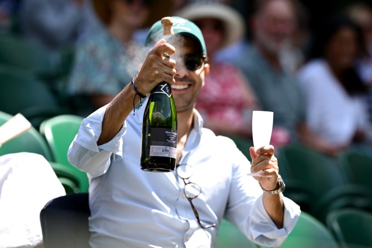 Man in sunglasses and cap spraying champagne, holding a flute at a live sport event, symbolizing high ticket prices.
