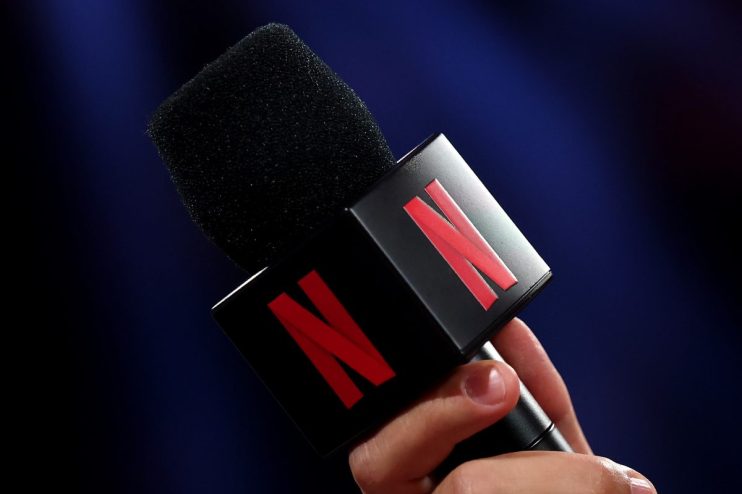 Hand holding a microphone with the Netflix logo on its cube, dark background.