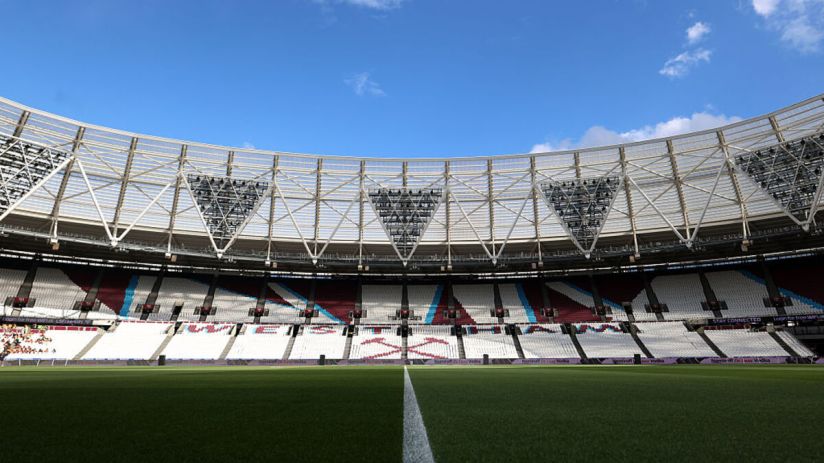 Empty stadium interior with green pitch, white sideline, and maroon, blue, and white seating under a clear blue sky.