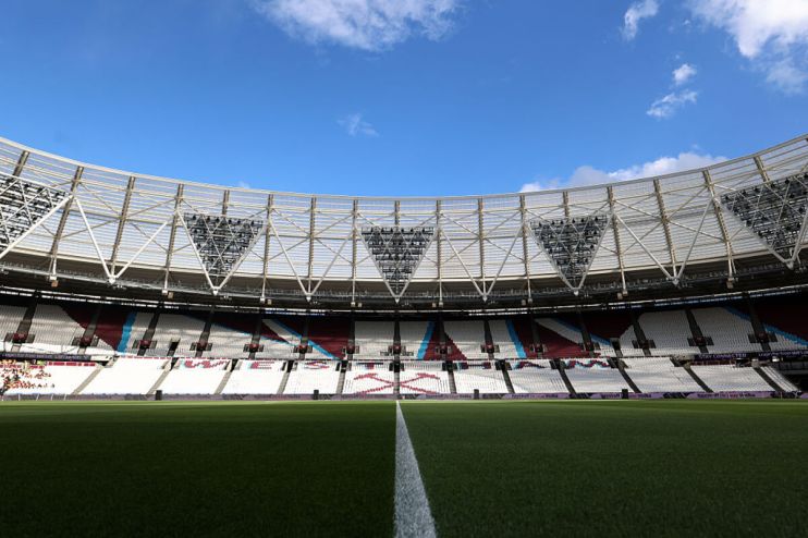 Empty stadium interior with green pitch, white sideline, and maroon, blue, and white seating under a clear blue sky.