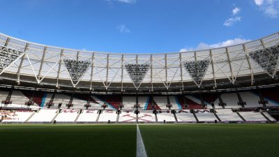 Empty London Stadium interior with green pitch, white sideline, and maroon/blue seating under a blue sky.