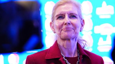 An older woman with blonde hair in a red jacket and silver necklace looks forward, smiling slightly.