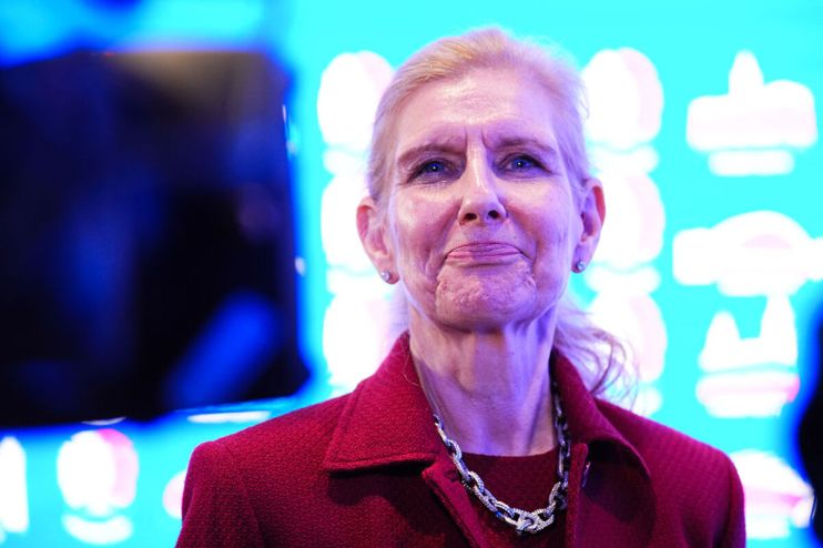 An older woman with blonde hair in a red jacket and silver necklace looks forward, smiling slightly.