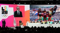 Olympic Games LA28 presentation with speaker and athlete images on screens, officials seated below.