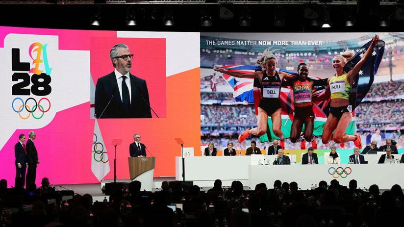 Olympic Games LA28 presentation with speaker and athlete images on screens, officials seated below.