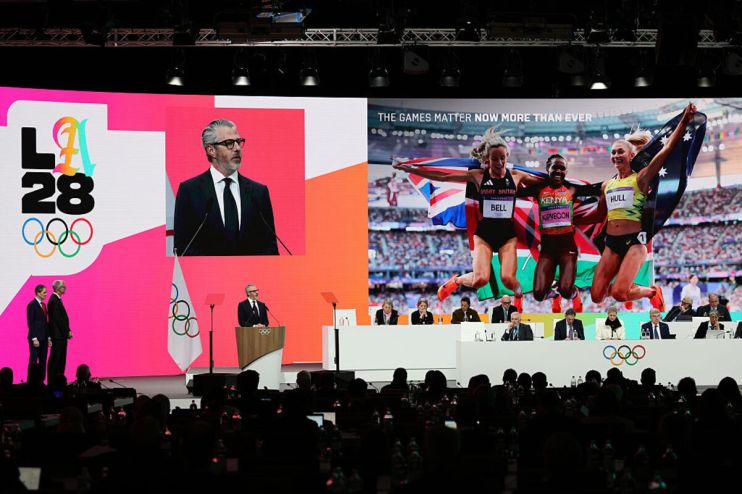 Olympic Games LA28 presentation with speaker and athlete images on screens, officials seated below.