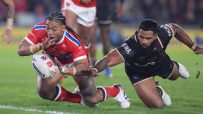 Two rugby players on green grass. One in red dives for a white ball, the other in dark jersey tackles.