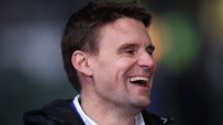 Close-up of a man laughing, looking right, with a dark jacket and white collared shirt
