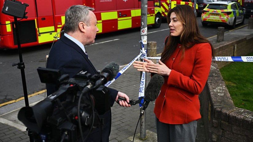 Journalist in red jacket interviewing a man near police and fire vehicles, with Police Line Do Not Cross tape.