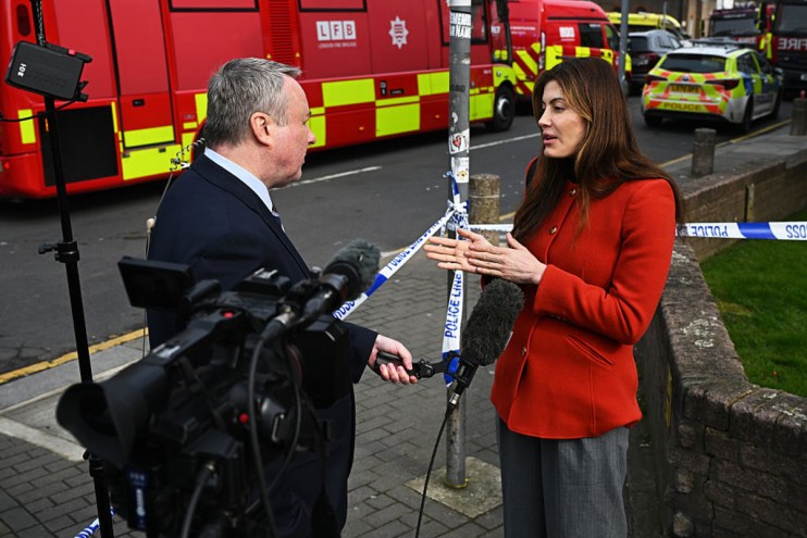 Journalist in red jacket interviewing a man near police and fire vehicles, with Police Line Do Not Cross tape.