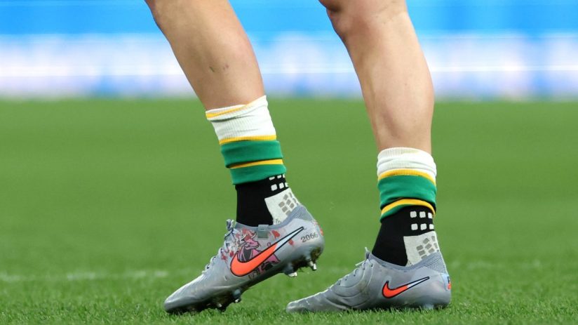 Athletes legs and feet in grey Nike cleats with orange swoosh, green and yellow socks on a green sports field.