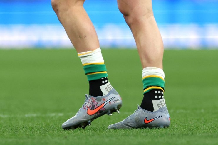 Athletes legs and feet in grey Nike cleats with orange swoosh, green and yellow socks on a green sports field.
