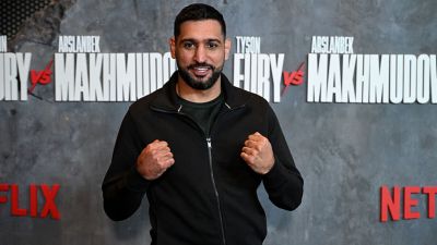 Amir Khan, former boxing champion, clenches fists in front of a Fury vs Makhmudov promotional backdrop