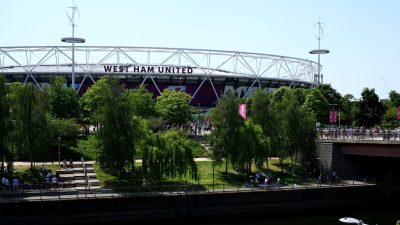 West Ham United stadium with its white structural roof against a clear sky, surrounded by green trees and a river.