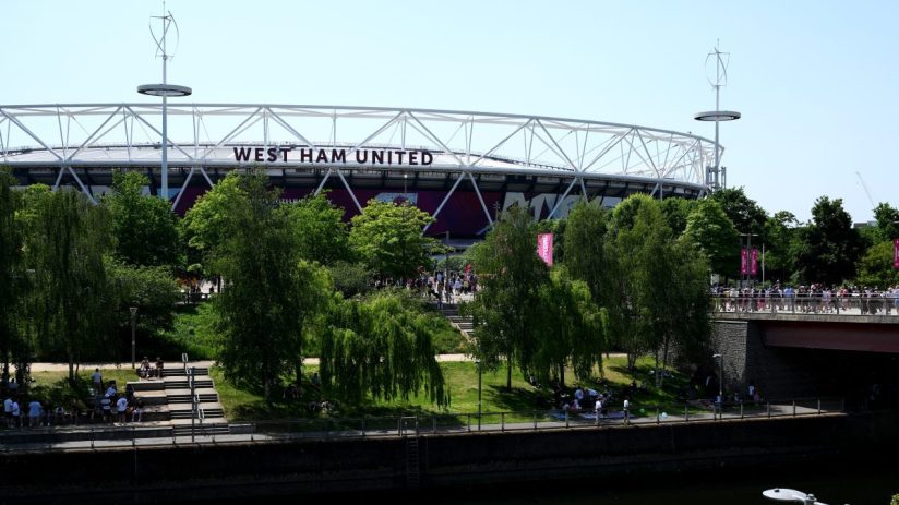 West Ham United stadium with its white structural roof against a clear sky, surrounded by green trees and a river.