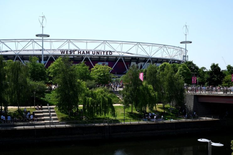 West Ham United stadium with its white structural roof against a clear sky, surrounded by green trees and a river.