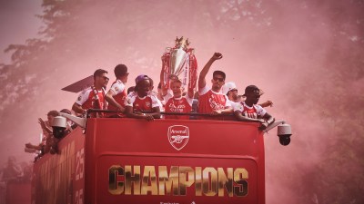 Arsenal team on a red double-decker bus parade, celebrating with the Premier League trophy amidst red smoke.