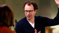 A man with curly hair and blue glasses gestures animatedly while speaking to an unseen individual.