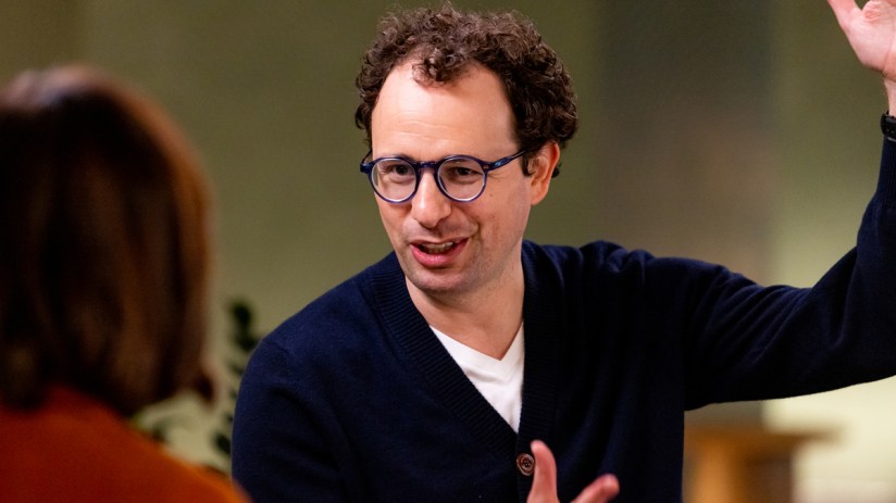A man with curly hair and blue glasses gestures animatedly while speaking to an unseen individual.