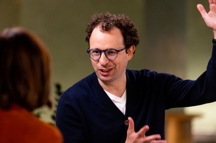 A man with curly hair and blue glasses gestures animatedly while speaking to an unseen individual.