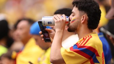Male fan in a yellow, red, and blue jersey drinks from a can, with a flag painted on his cheek, holding a phone.