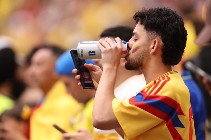 Male fan in a yellow, red, and blue jersey drinks from a can, with a flag painted on his cheek, holding a phone.