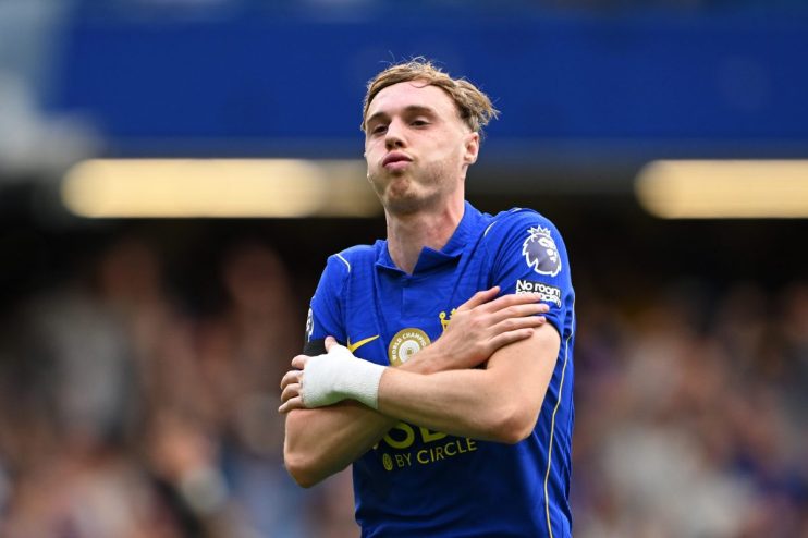 Chelsea footballer Conor Gallagher in a blue jersey, with a white wrist wrap, arms crossed on the field.