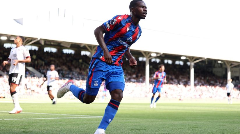 Footballer in a blue and red jersey running on a green field during a sunny match.