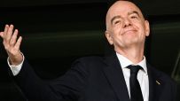 Gianni Infantino, FIFA President, smiling and waving, wearing a dark suit and tie.
