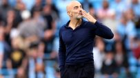 Bald man with a beard in a navy polo shirt blowing a kiss to a crowd at a stadium