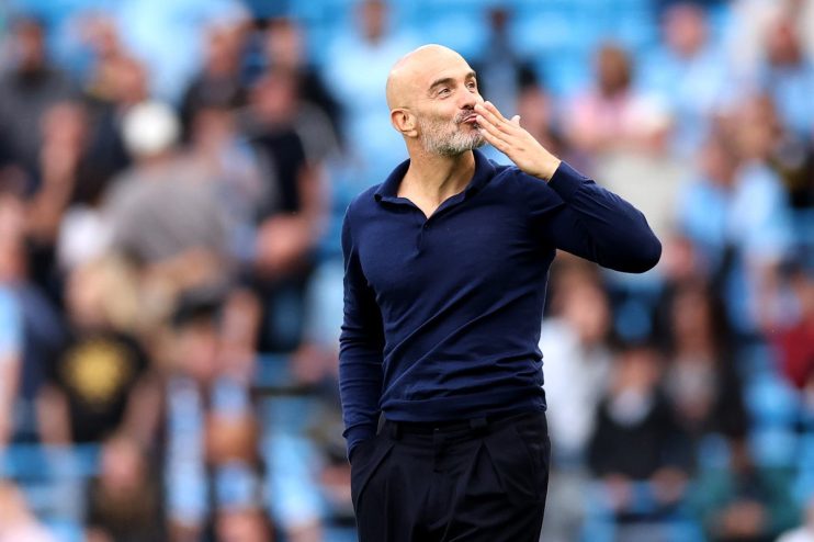 Bald man with a beard in a navy polo shirt blowing a kiss to a crowd at a stadium