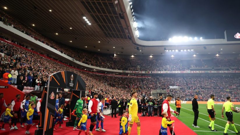 Football players and child mascots enter a brightly lit stadium packed with cheering fans at night.