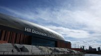 Hill Dickinson Stadium exterior, with modern architecture, waterfront access, and distant city skyline under a cloudy sky.