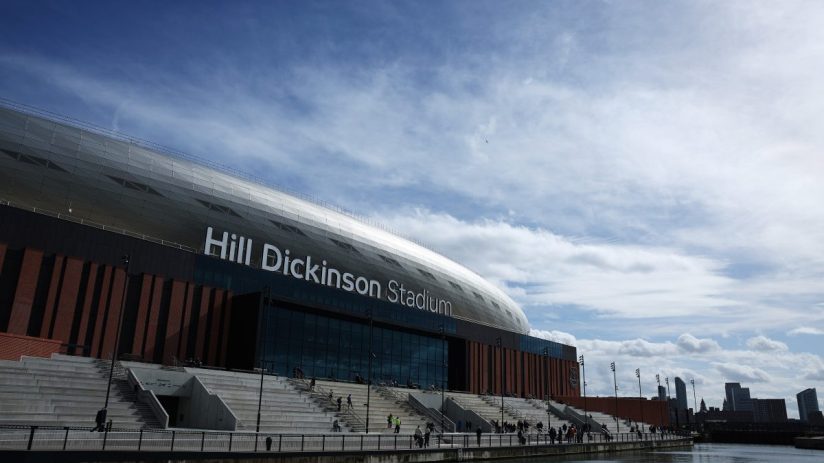 Hill Dickinson Stadium exterior, with modern architecture, waterfront access, and distant city skyline under a cloudy sky.