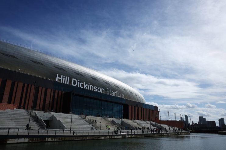 Hill Dickinson Stadium exterior, with modern architecture, waterfront access, and distant city skyline under a cloudy sky.