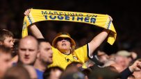 Female football fan in yellow hat and jersey holding a Marching On Together scarf overhead in a stadium crowd.
