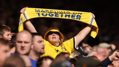 Female football fan in yellow hat and jersey holding a Marching On Together scarf overhead in a stadium crowd.