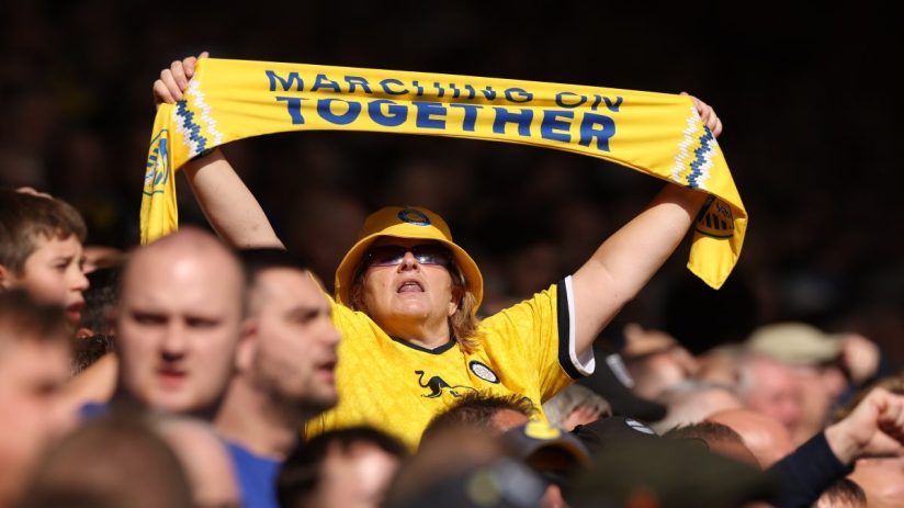 Female football fan in yellow hat and jersey holding a Marching On Together scarf overhead in a stadium crowd.