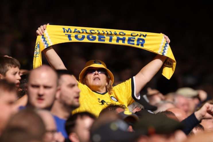 Female football fan in yellow hat and jersey holding a Marching On Together scarf overhead in a stadium crowd.