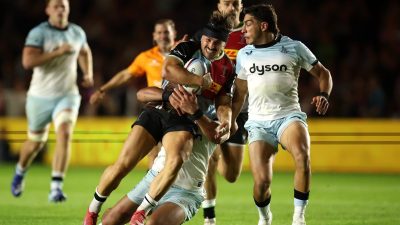 Rugby player in brown and red jersey being tackled by opponent in light blue jersey on green field