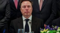 Elon Musk in a dark suit and blue tie, seated at a table with a glass and microphone.